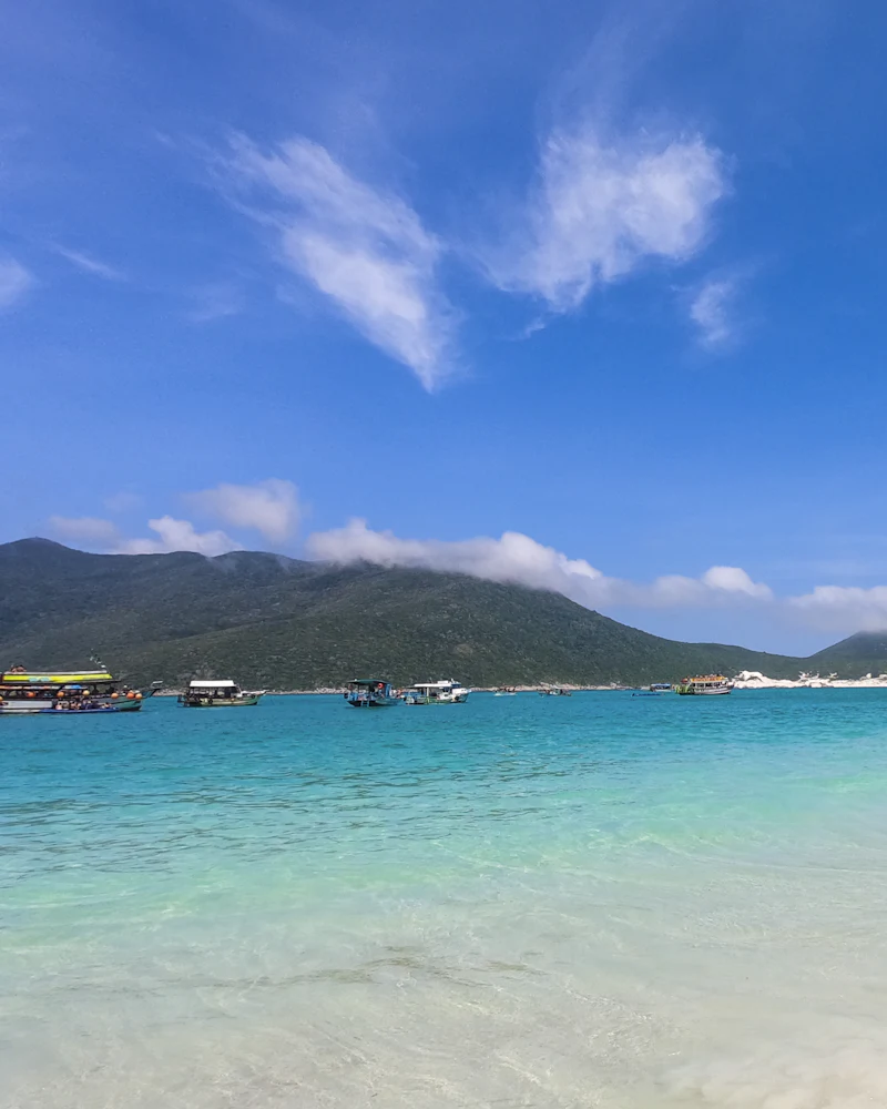 Água clara e azul-turquesa na beira da praia, com barcos e morros ao fundo