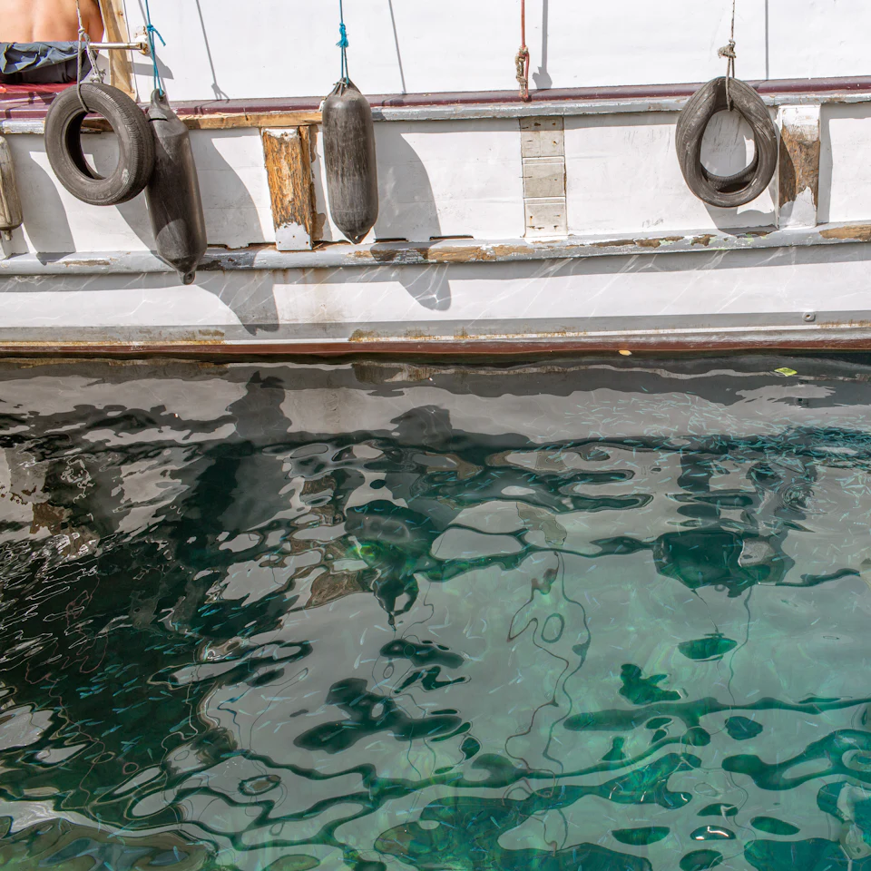 Costado de um barco branco com defensas penduradas sobre água cristalina