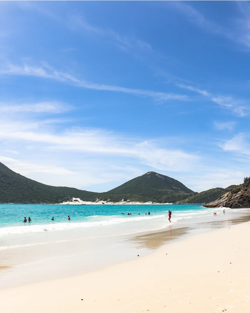 Praia de areia clara e mar turquesa, com morros ao fundo e banhistas na água